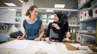 German seamstress works with a trainee Syrian refugee at a fashion workshop