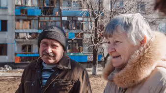 An older man and woman stand in front of an apartment building